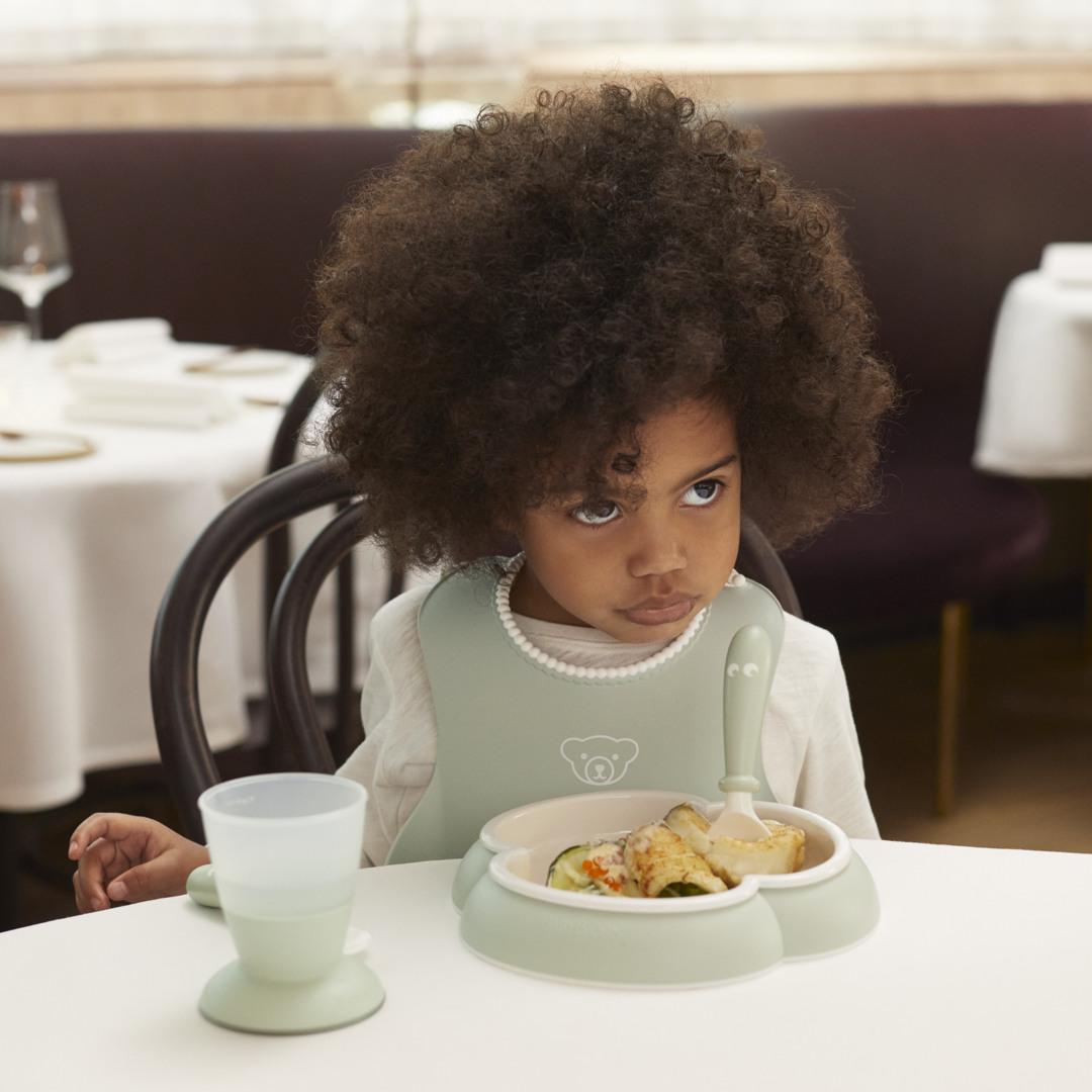 Young child with curly hair, wearing a bib, sitting at a table with a meal and a cup in front of them, looking to the side.