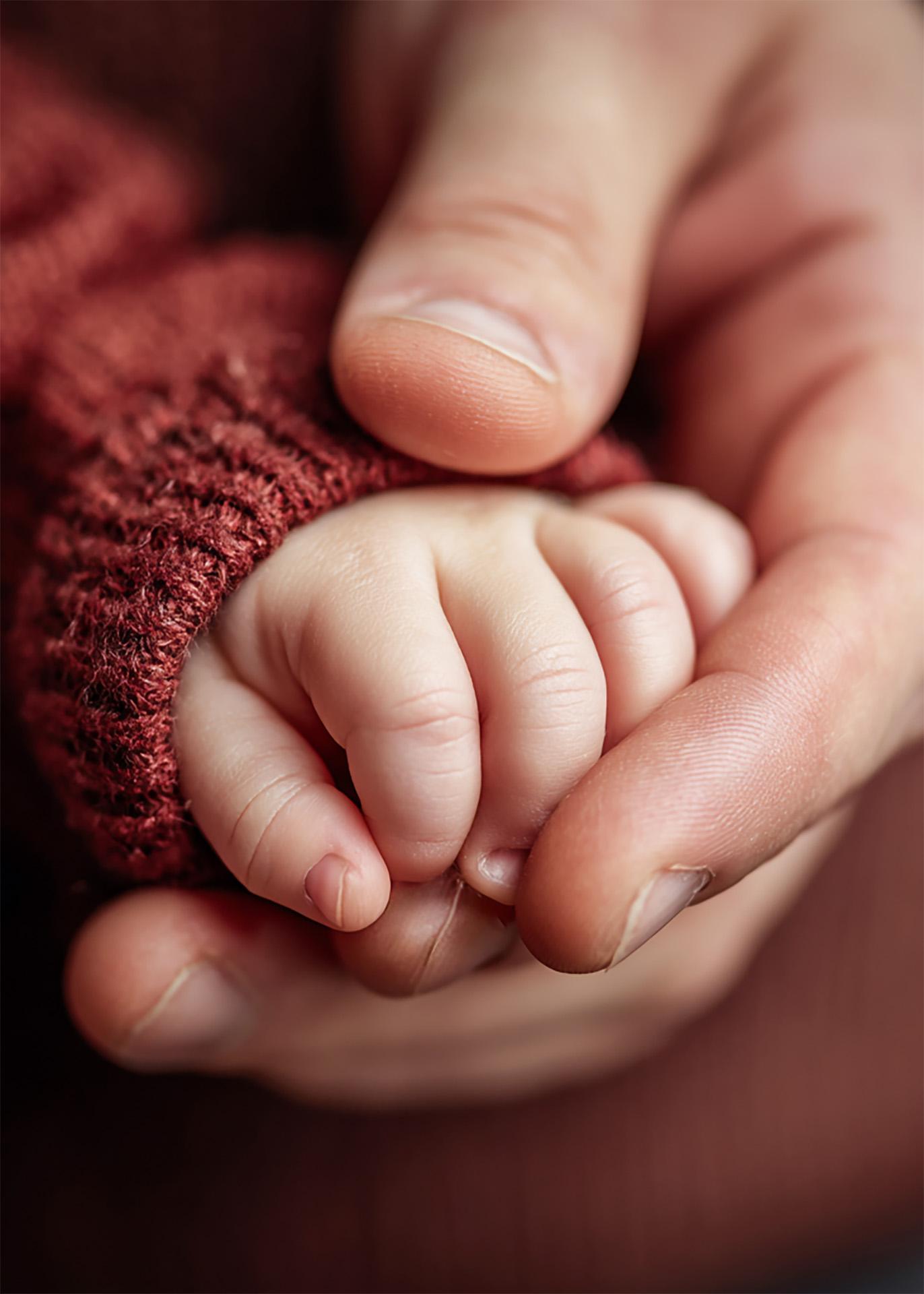 Close-up of an adult hand gently holding a newborn's tiny hand in a red knit sleeve.