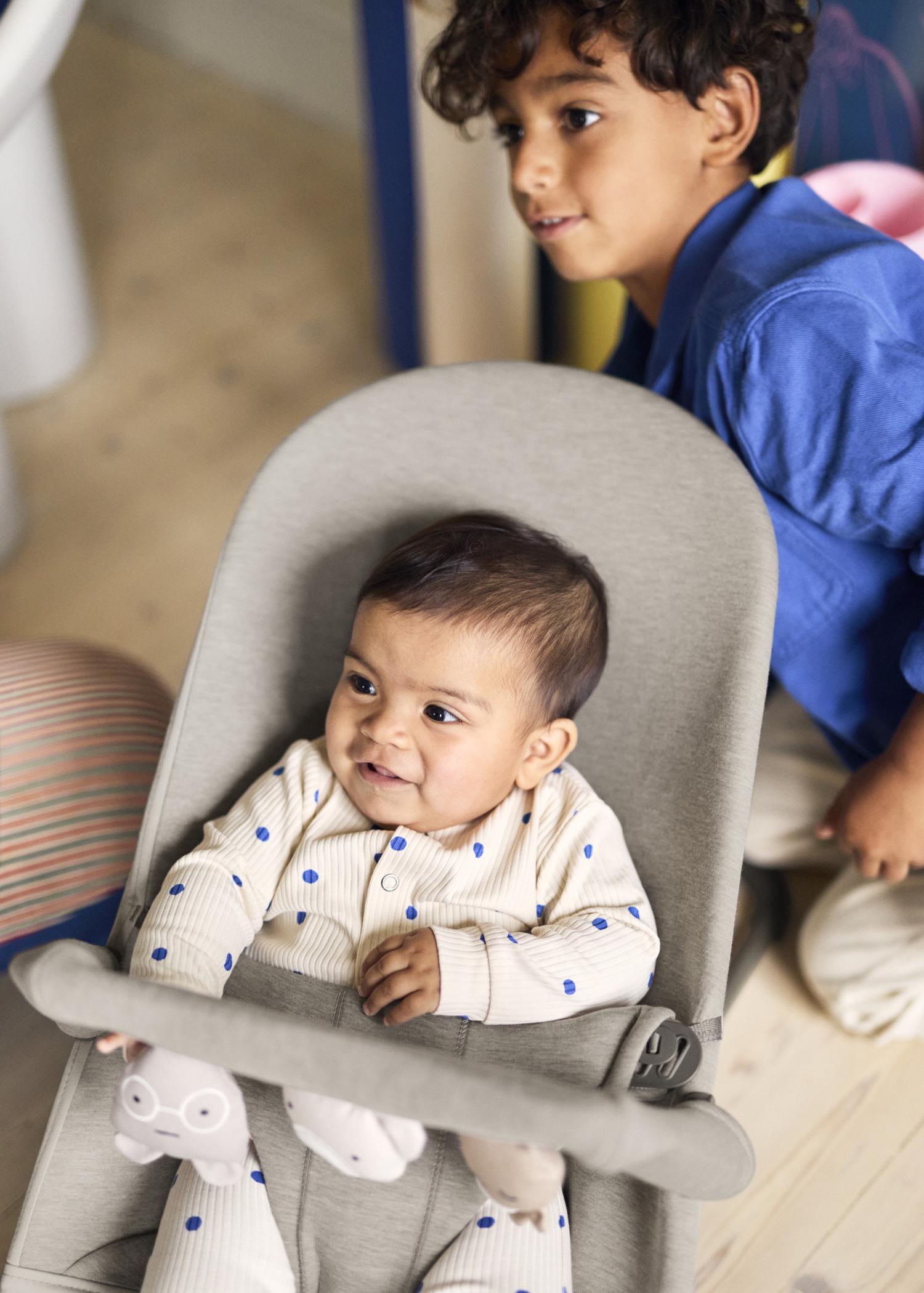 A baby in a polka dot outfit sits in a baby bouncer, while a young child in a blue shirt stands nearby, both smiling.