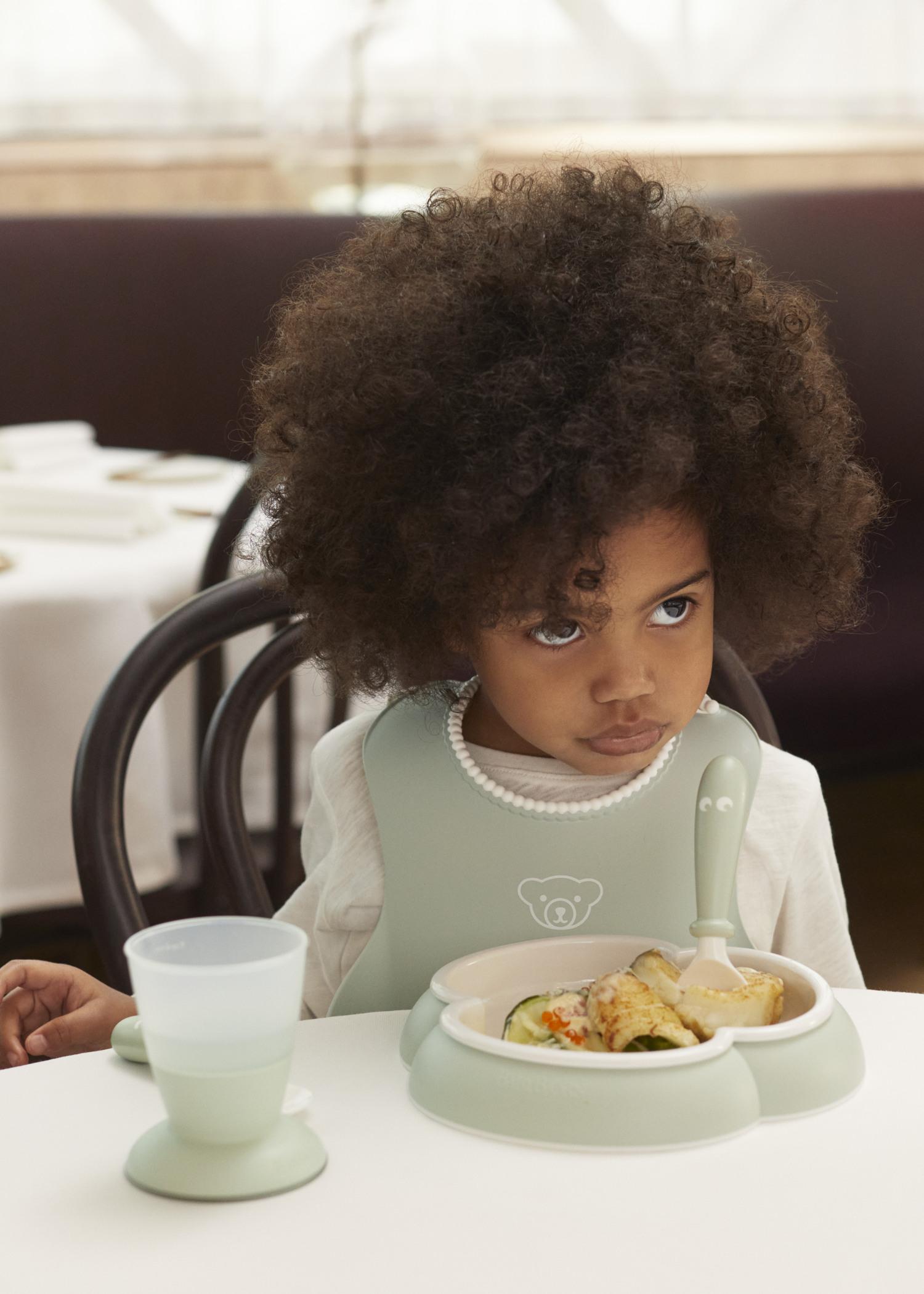 Young child with curly hair, wearing a bib, sitting at a table with a meal and a cup in front of them, looking to the side.