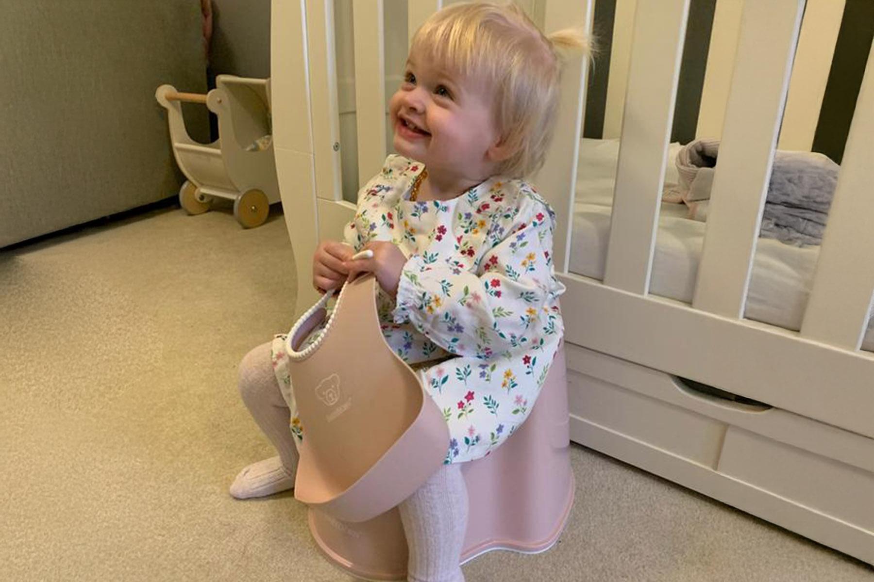 Smiling toddler in floral pajamas sits on a pink potty in a nursery, holding a silicone bib beside a white crib.