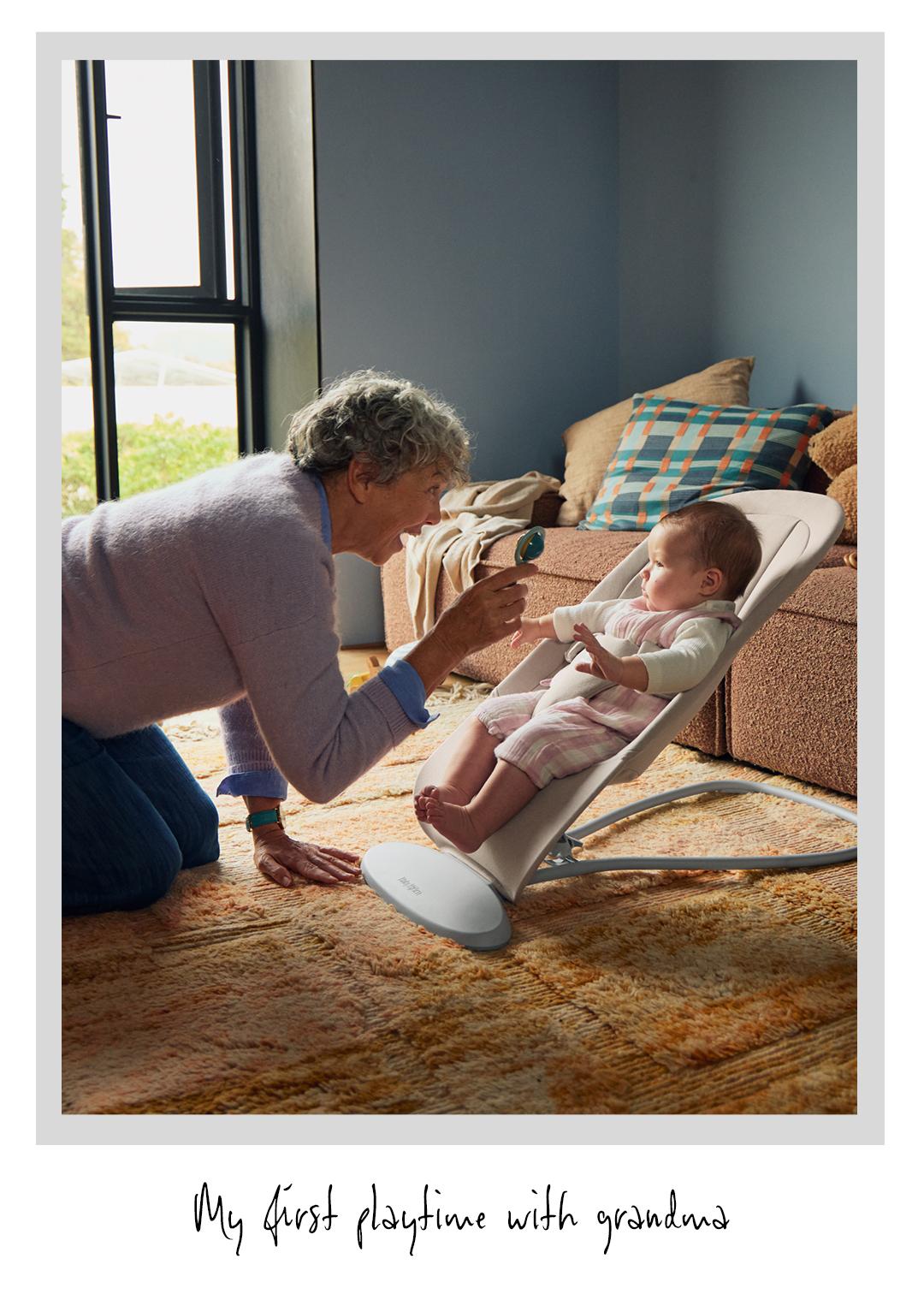 Elderly woman kneeling and playing with a baby in a bouncer chair at home, with a cozy living room setting.