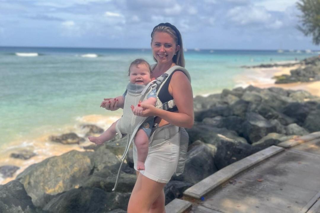 A woman smiling while carrying a baby in a harness, standing by a rocky shoreline with the ocean and blue sky in the background.