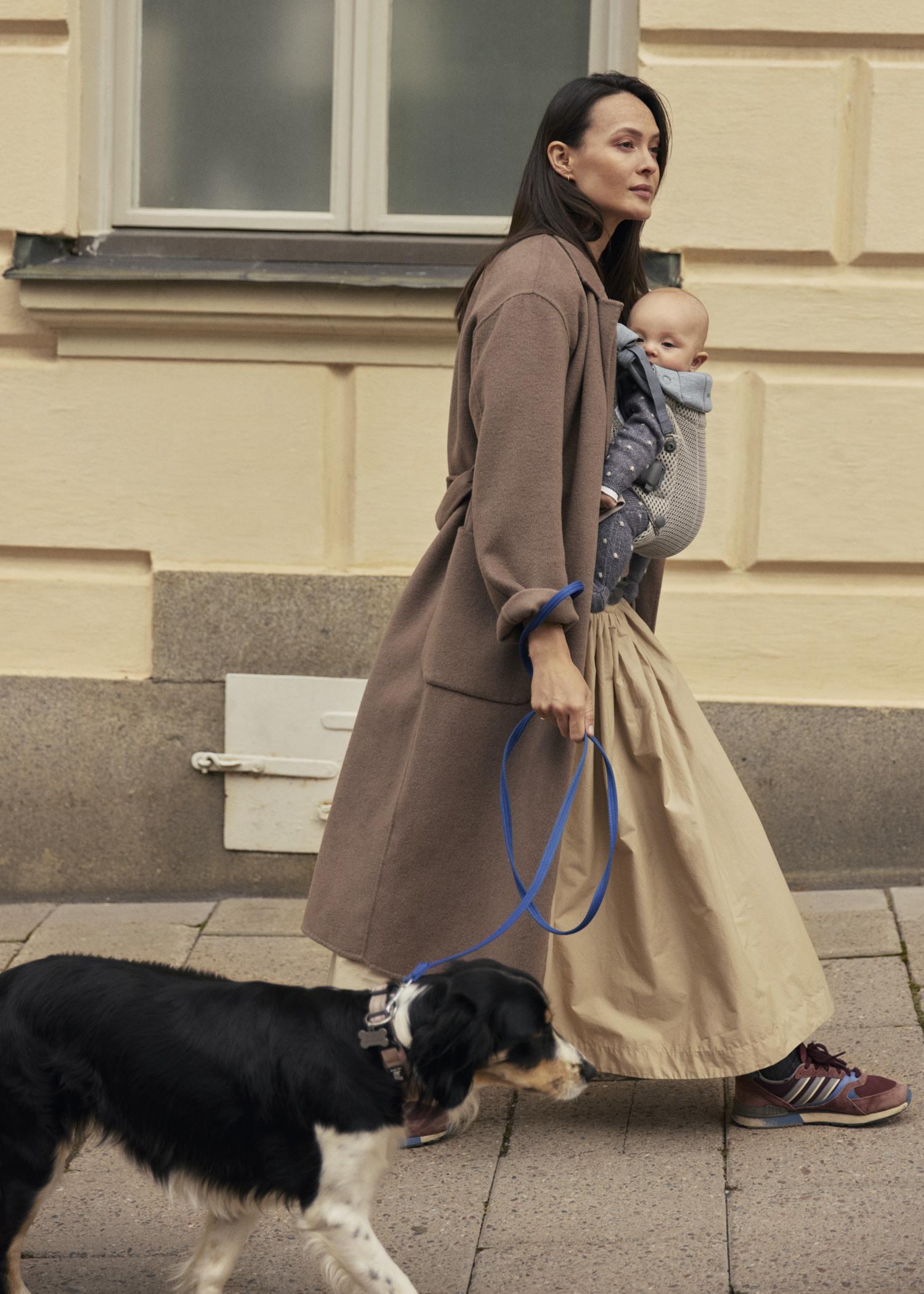 Woman walking on sidewalk with a baby in a beige carrier and a dog on a leash. She wears a brown coat and beige skirt.