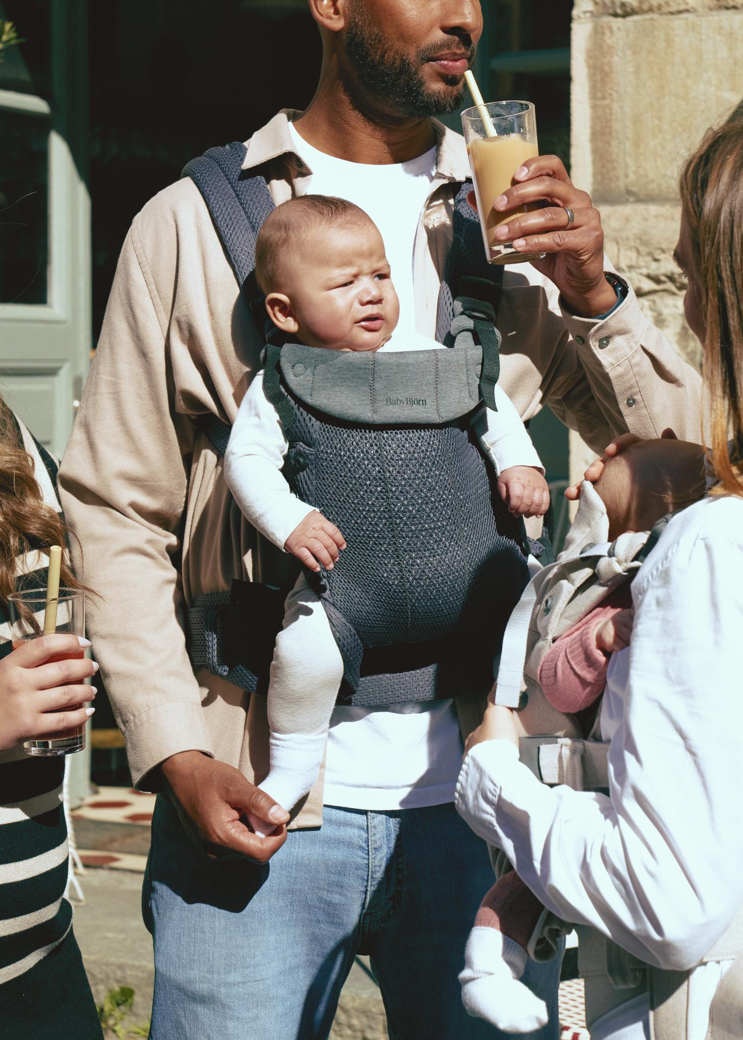 Man holding a baby in a carrier, drinking a smoothie outdoors. The baby is wearing a white outfit and appears to be squinting in the sunlight.