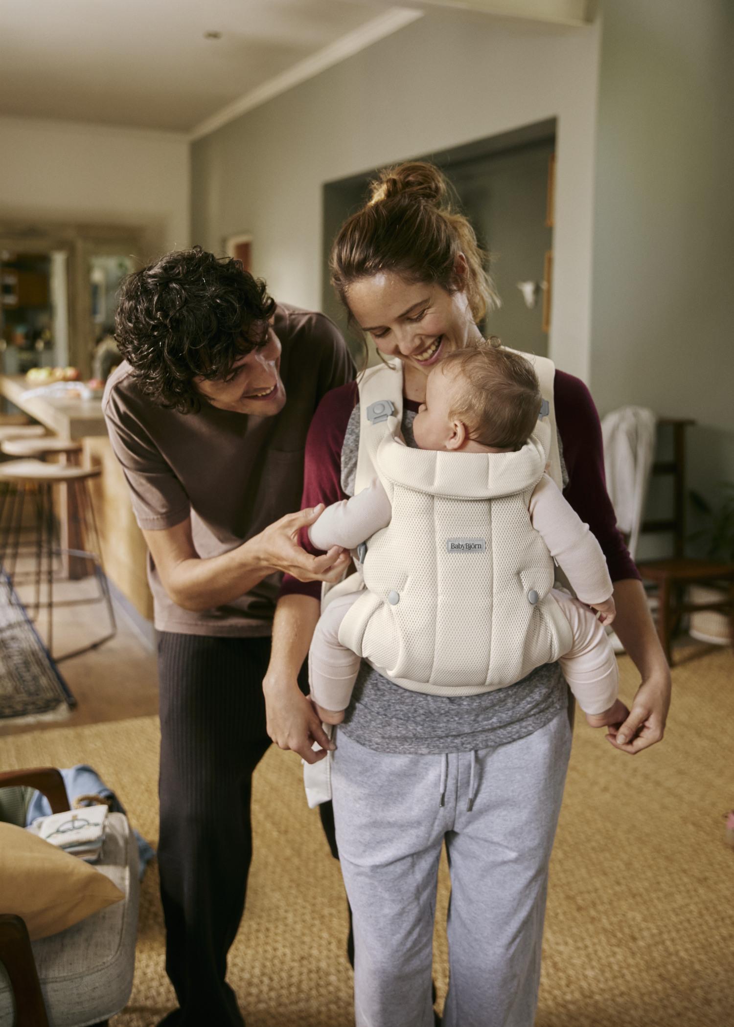 A smiling woman carries a baby in a white carrier, while a man leans in, touching the baby's hand, in a cozy home setting.