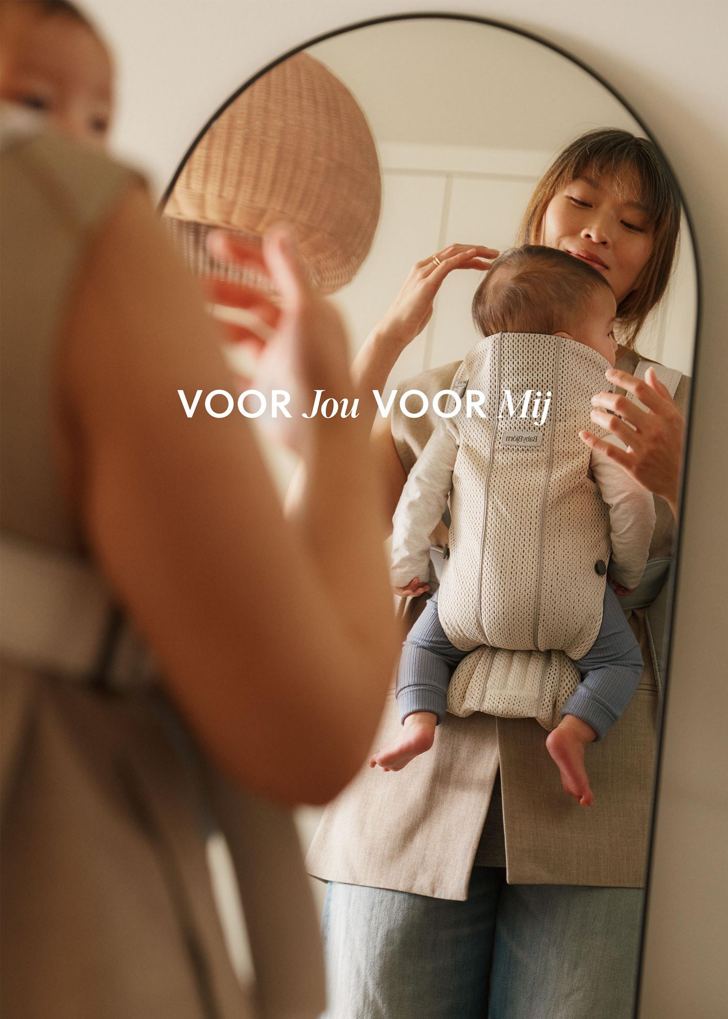 Woman holding a baby in a carrier, looking in a mirror. Text on the mirror reads "VOOR Jou VOOR Mij."