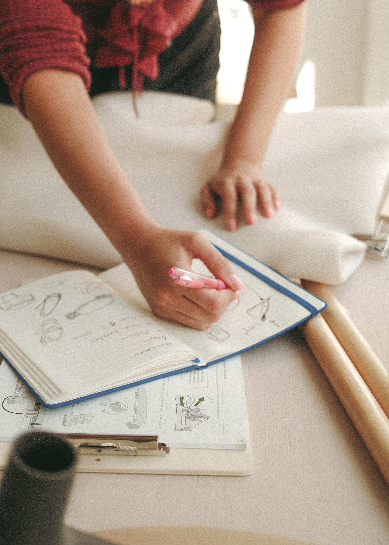 Persoon tekent schetsen in een notitieboekje met een roze pen, op een tafel bedekt met papieren en stoffen.