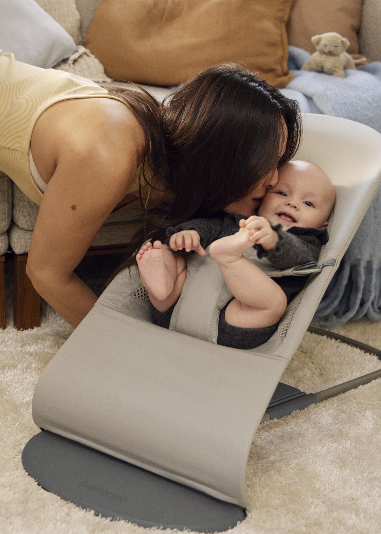 A woman gently kisses a smiling baby lying in a gray baby bouncer on a soft carpet, with a plush toy in the background.