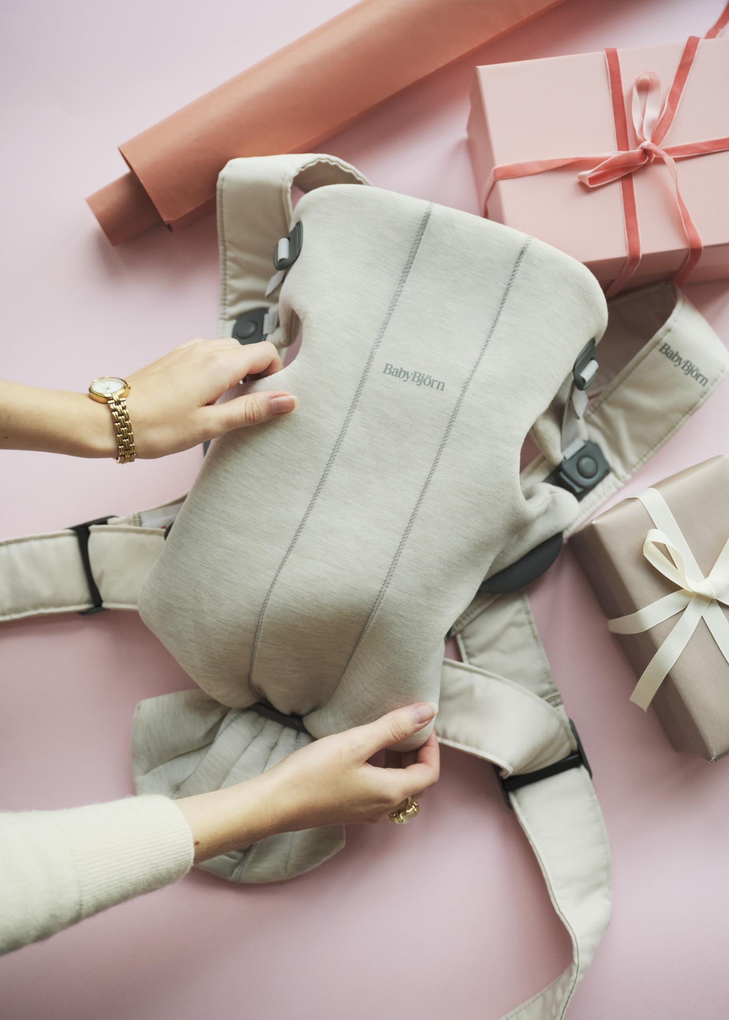 Hands holding a beige baby carrier on a pink surface, surrounded by pink gift boxes and wrapping paper.