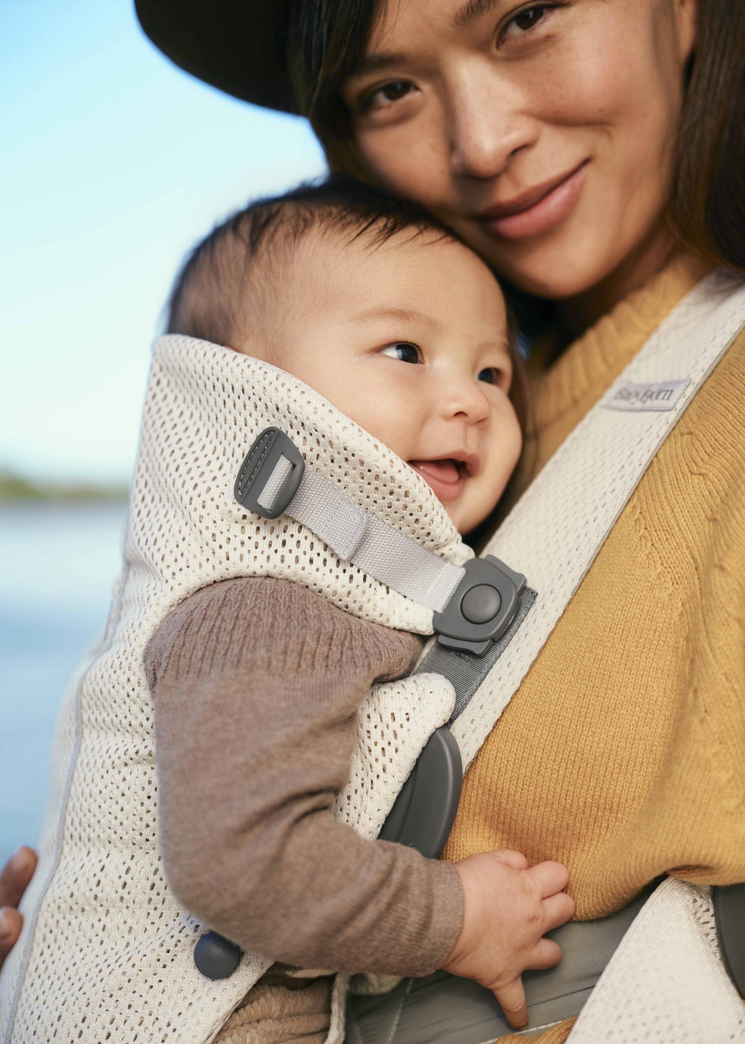 A smiling woman holds a happy baby in a beige baby carrier outdoors, with blue sky in the background.