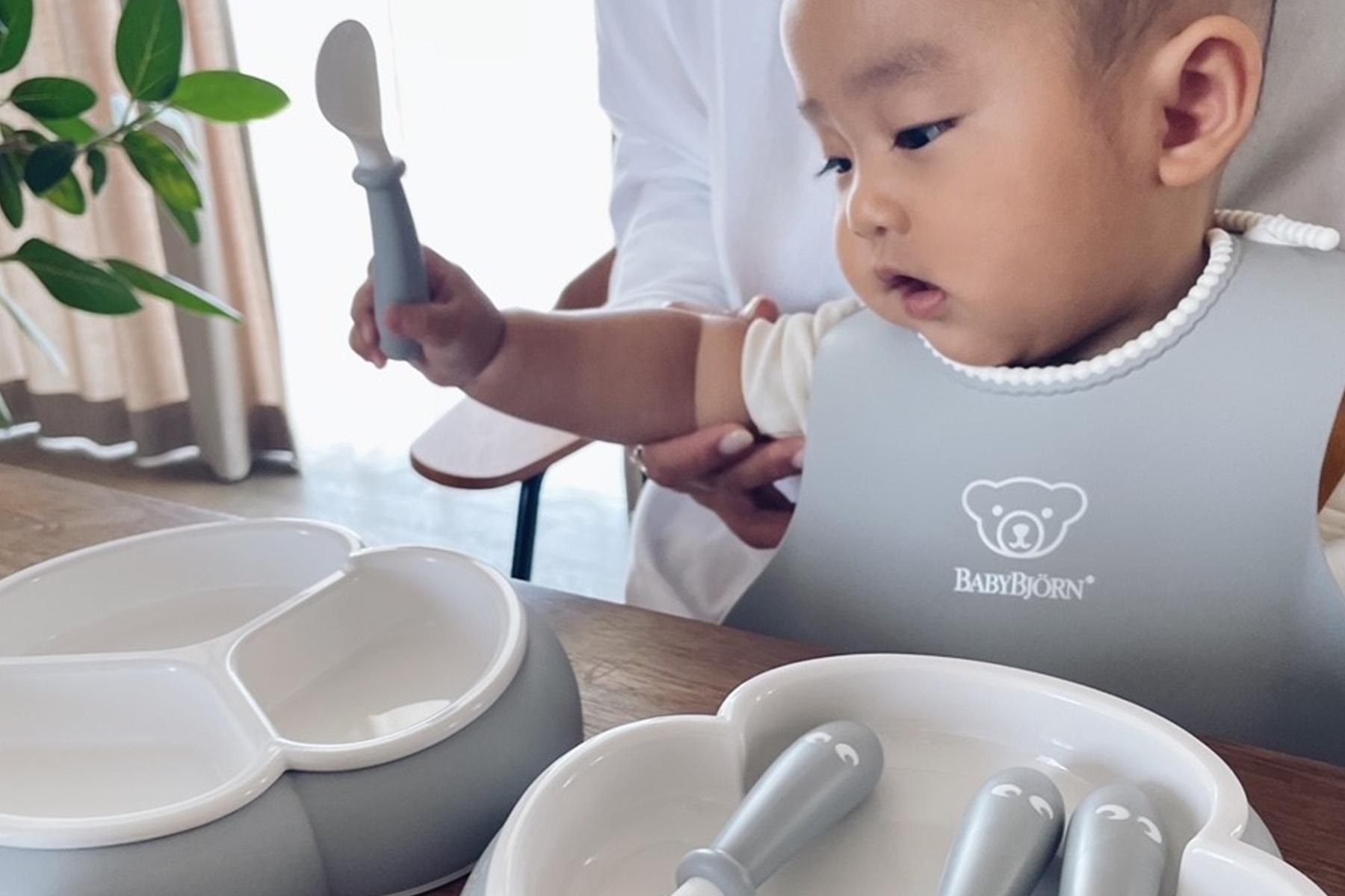 A baby in a bib holds a spoon, reaching towards a set of divided plates and utensils on a table. A person supports the baby.