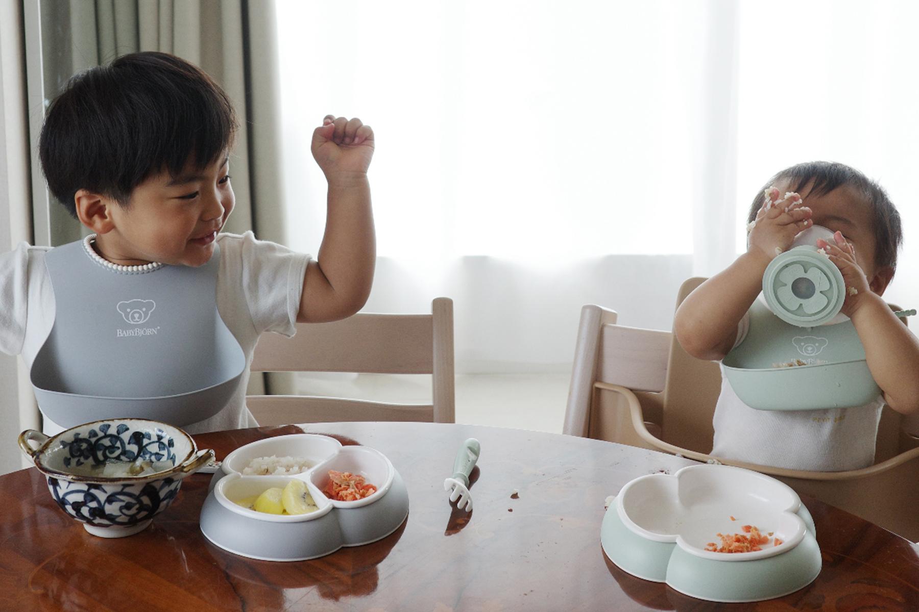 Two toddlers in high chairs enjoying a meal. One child raises a hand excitedly, while the other drinks from a bottle.