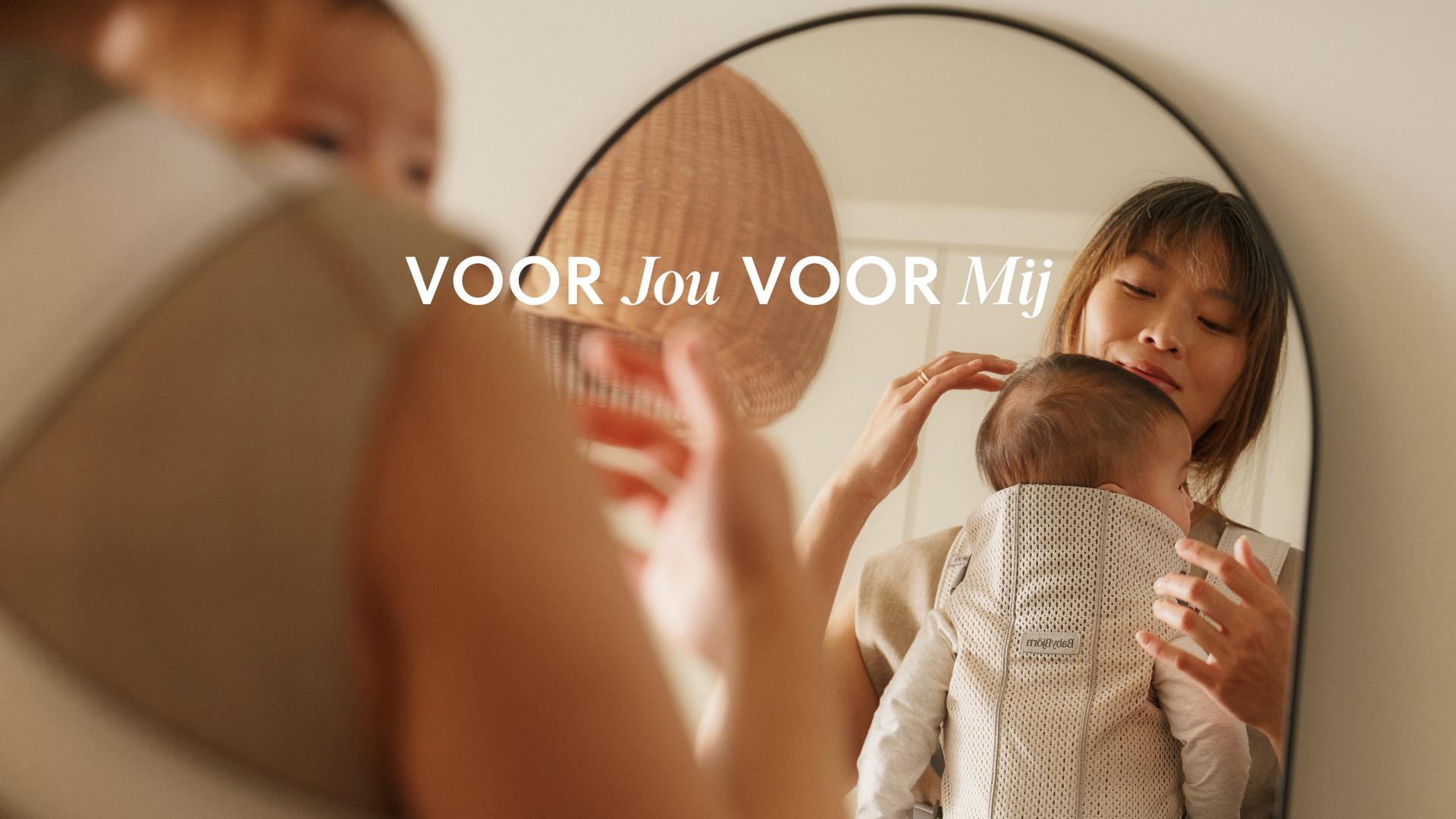 A woman holding a baby in a carrier is reflected in a mirror. Text reads "VOOR Jou VOOR Mij." A woven basket hangs nearby.