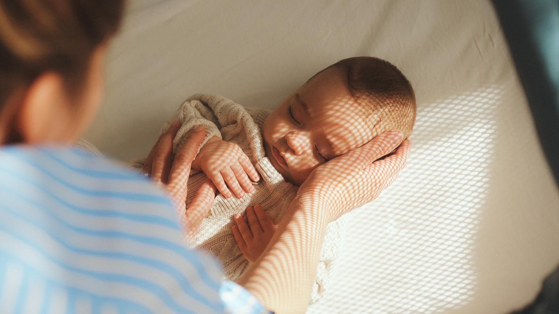 A person gently cradles a sleeping baby lying down, with soft sunlight creating patterns on the baby's face.