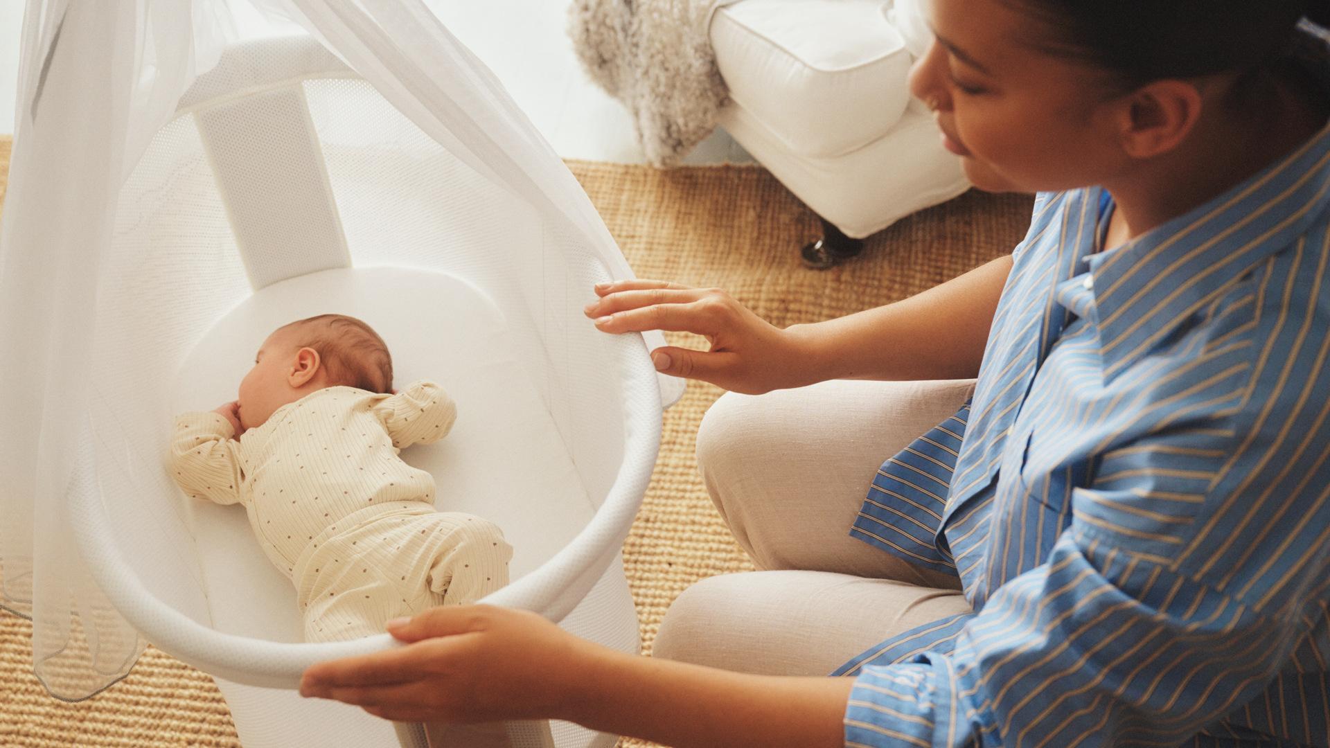 A woman in a striped shirt gently touches a cradle with a sleeping baby in a beige onesie. The setting is calm and nurturing.