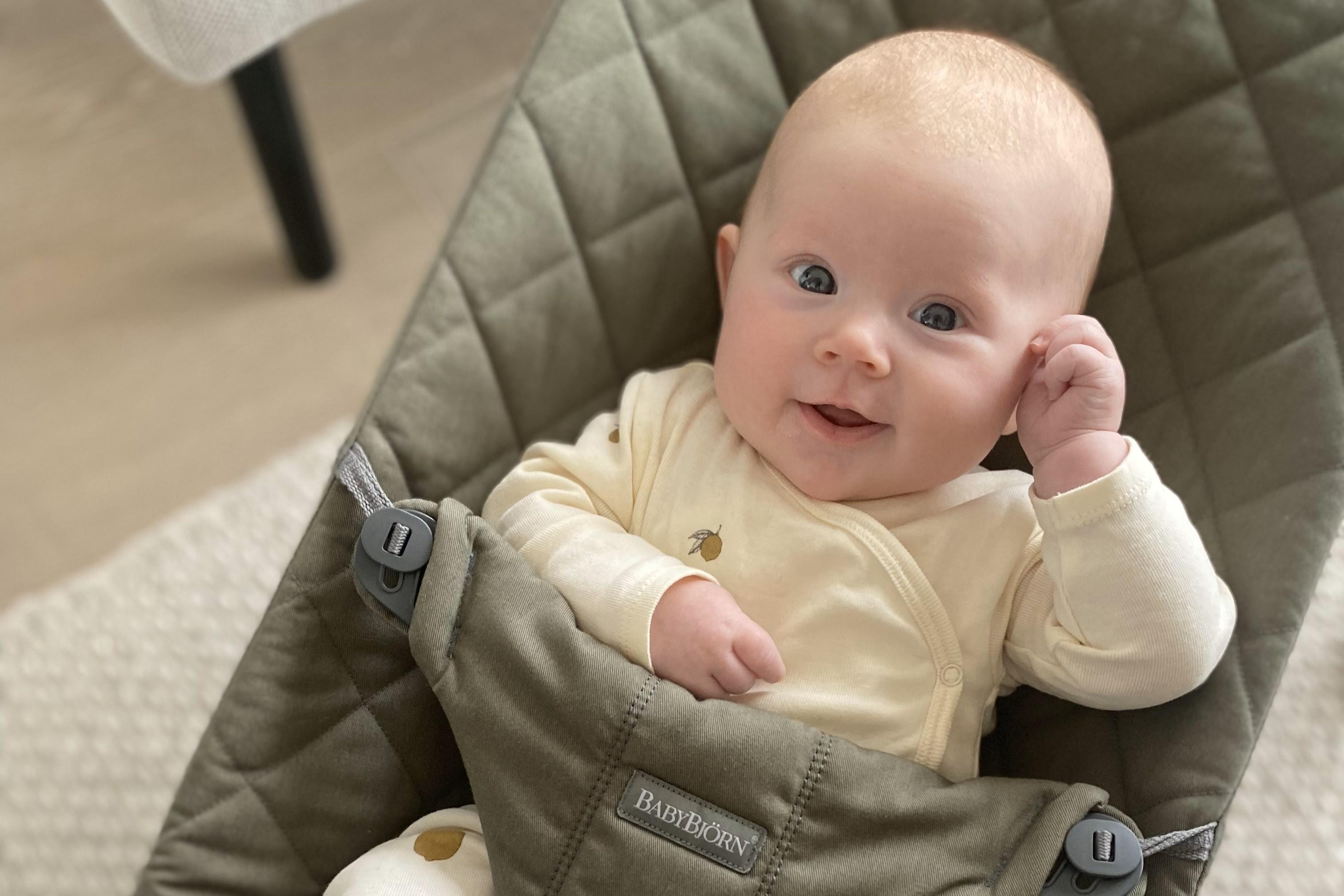 A baby in a white onesie sits in a green quilted chair, smiling and looking up with one hand near their ear.