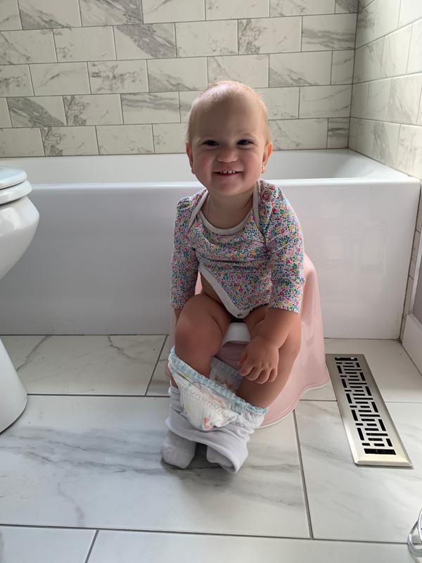 A smiling baby sits on a pink potty in a tiled bathroom, with a diaper around their ankles, in front of a bathtub.
